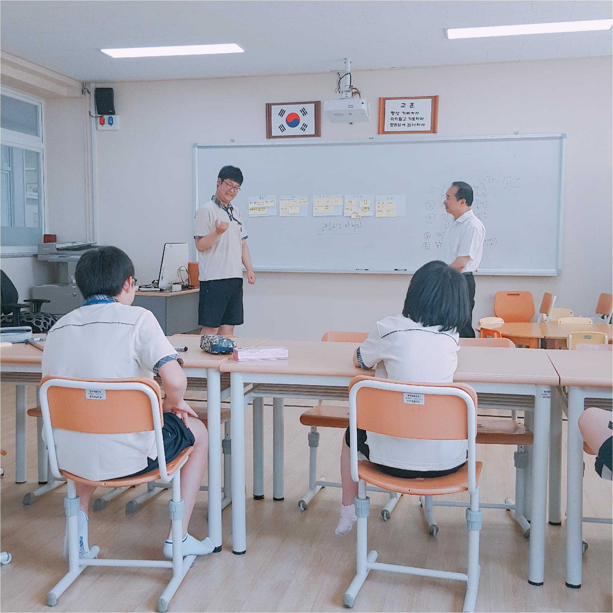 한 학생이 발표를 하고 있고, 선생님과 나머지 학생들은 발표하는 학생을 보고 있습니다.