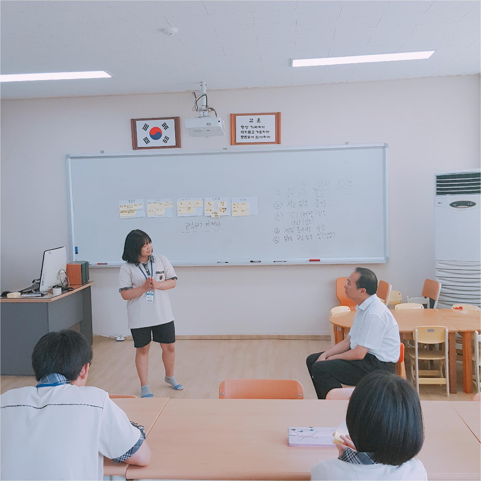 한 학생이 발표를 하고 있고, 선생님과 나머지 학생들은 발표하는 학생을 보고 있습니다.