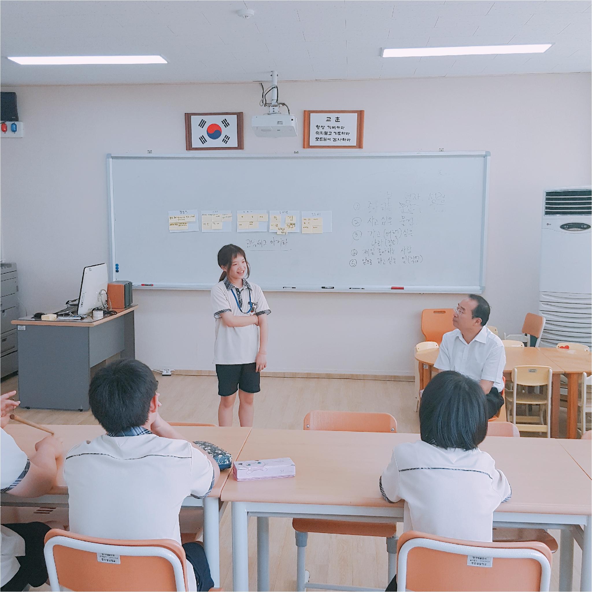 한 학생이 발표를 하고 있고, 선생님과 나머지 학생들은 발표하는 학생을 보고 있습니다.